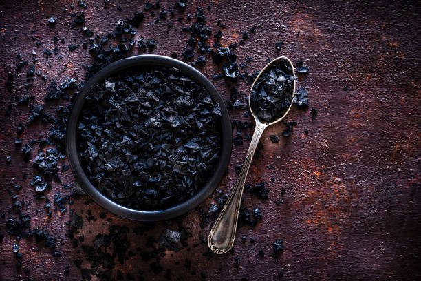 Top view of a black bowl filled Hawaiian salt shot on abstract brown rustic table. A vintage metal spoon with salt is beside the bowl and Hawaiian salt crystals are scattered on the table. Useful copy space available for text and/or logo. Predominant colors are brown and black. Low key DSRL studio photo taken with Canon EOS 5D Mk II and Canon EF 100mm f/2.8L Macro IS USM.