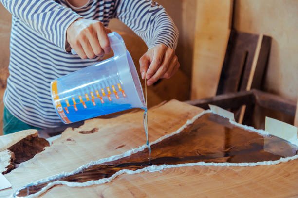 Closeup of carpenter pouring epoxy liquid in a wooden table. Process of making a craft resin and wood table